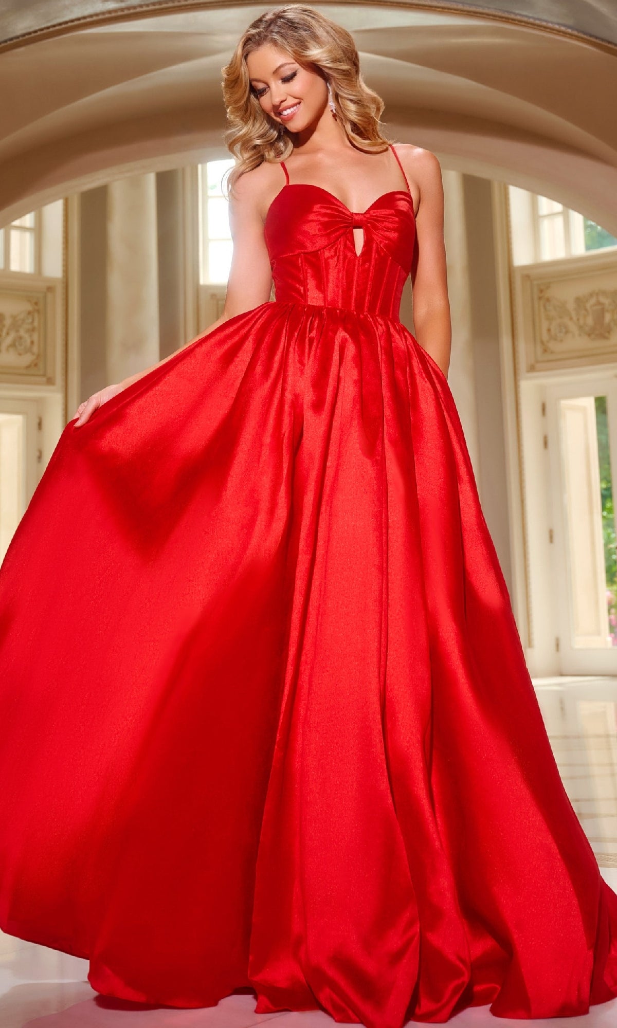 A woman stands indoors, holding out one side of her bright red Satin Prom Ball Gown (Jovani 43199), featuring a floor-length full skirt and fitted corset bodice.